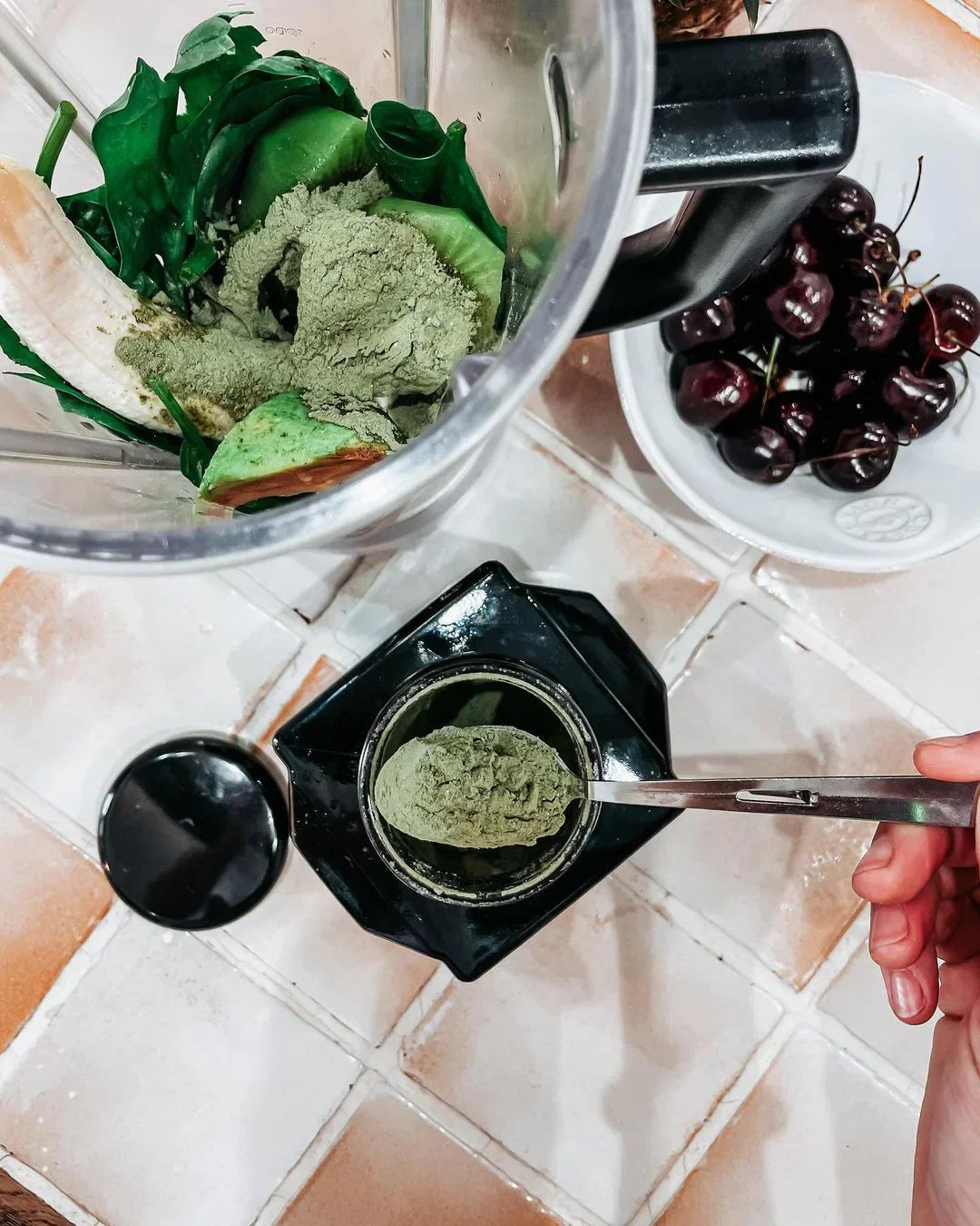 Blender with green ingredients, measuring spoon, and bowl of cherries.